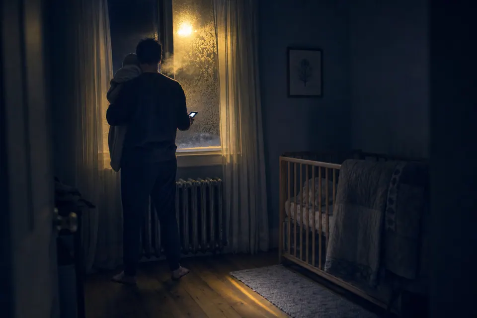 A family-housing nursery at night: a father seen from behind at the frosted window, his infant against his shoulder, a phone’s glow in his hand — the report being sent; the radiator below the window unlit.
